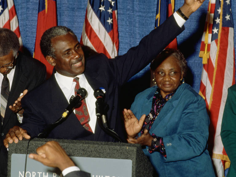 Color photo of North Carolina senatorial candidate Harvey Gantt speaking to supporters on election night in 1990, standing at a podium with his arm raised.