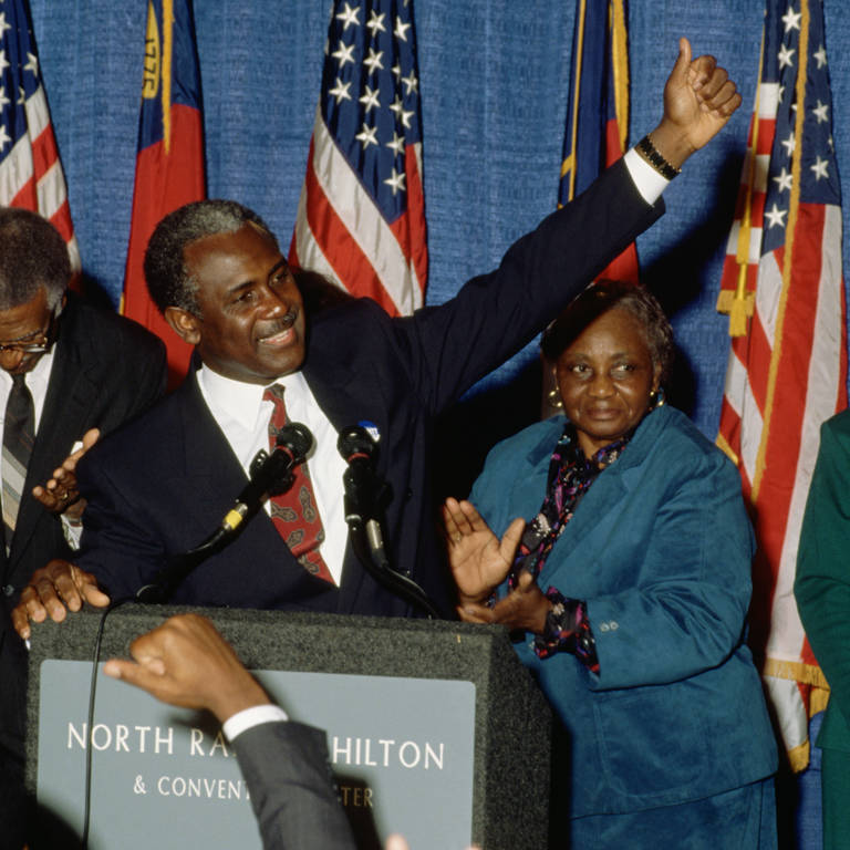 Color photo of North Carolina senatorial candidate Harvey Gantt speaking to supporters on election night in 1990, standing at a podium with his arm raised.