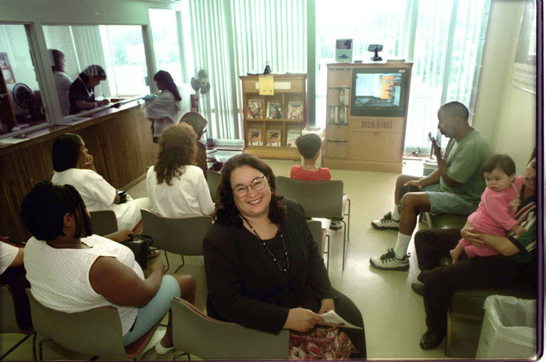 Color photo of Sylvia Drew Ivie, Executive Director of T.H.E. Clinic, sitting in a waiting room among the MediCal patients that the clinic serves.