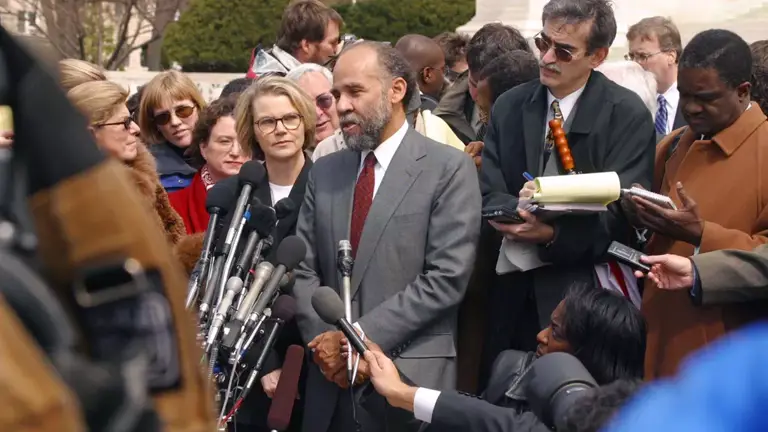 Color photo of John Payton speaking into a group of microphones outside, surrounded by reporters and other people.