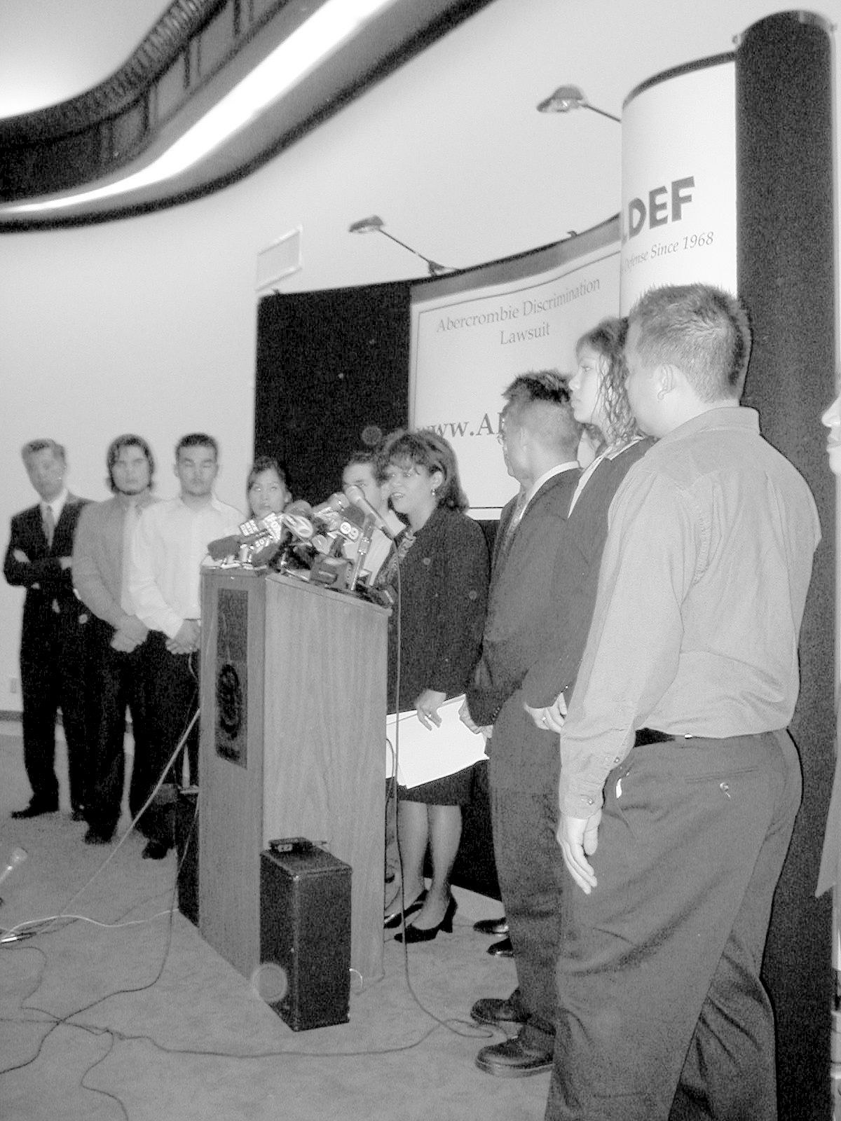 Black-and-white photo of several people standing in a conference room behind a podium with microphones. Behind them, a sign reads, "Abercrombie Discrimination Lawsuit."