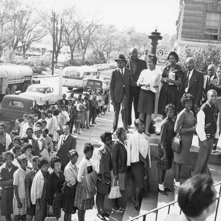 Black-and-white photo of a long line of Black people standing on the steps of a building and on the sidewalk.