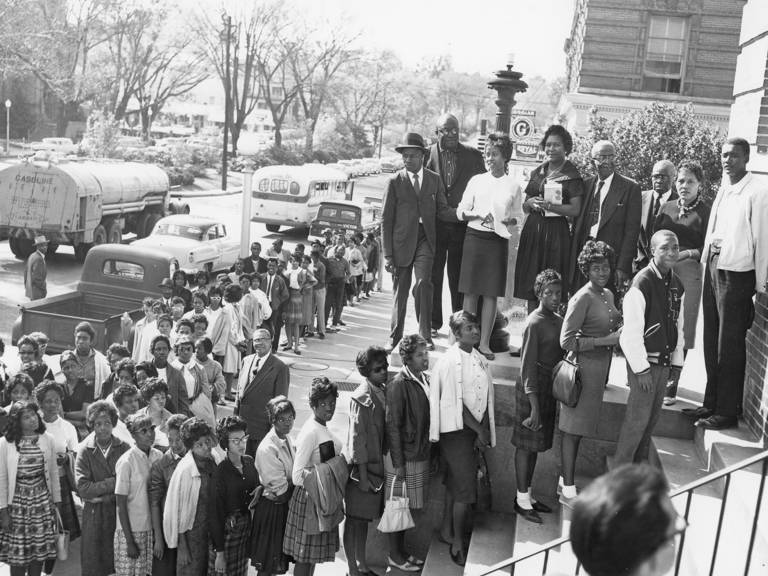 Black-and-white photo of a long line of Black people standing on the steps of a building and on the sidewalk.