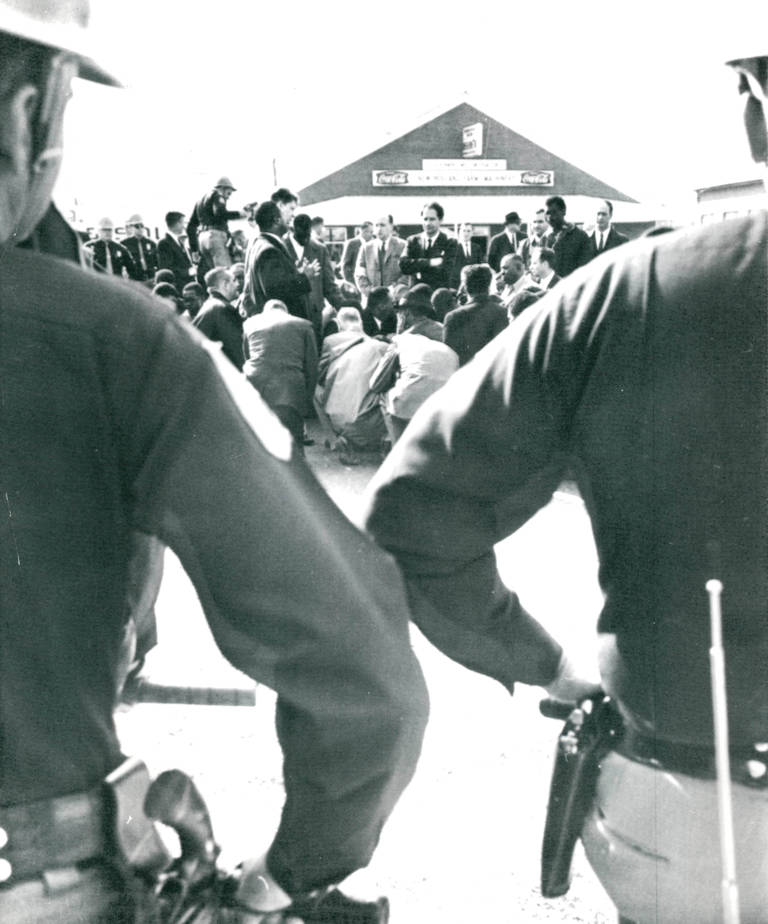 Black-and-white photo of state troopers standing by as demonstrators kneel and pray during a march.