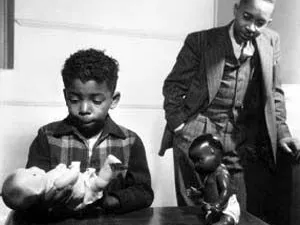 Black-and-white photo of a Black child seated at a table while holding a white doll, with a Black doll also on the table, as a Black man in a suit stands and observes him.