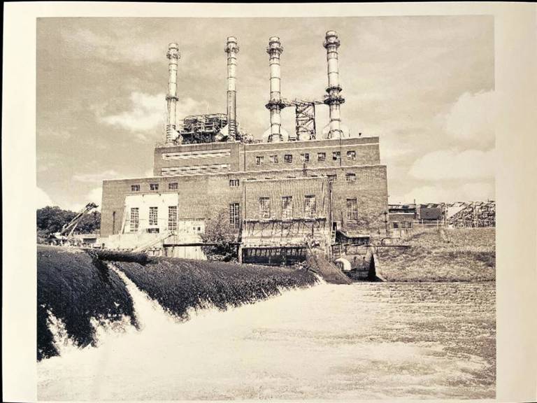 Photo of Duke Power Steam Station. In the foreground of the picture, water is flowing in front of the building. In the background is the Duke Power Steam Station, a two-story building with four steam pillars jutting out of the top.