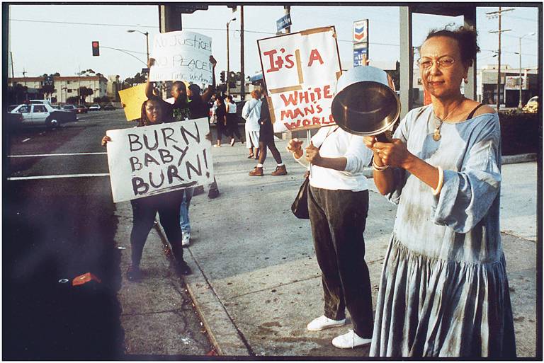 Color photo of a group of protesters standing on a sidewalk, holding pots and signs reading, "No justice, no peace," "Burn baby burn," and "It's a white man's world."