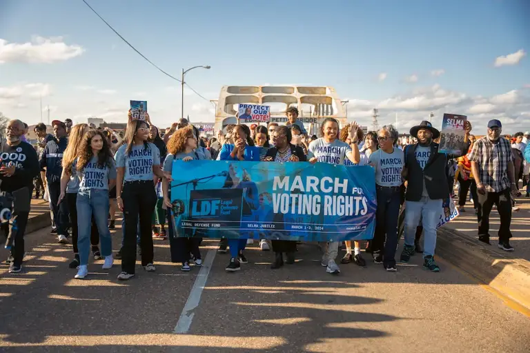 Color photo of LDF staff marching across the Edmund Pettus Bridge in Selma, Alabama, in March 2024 during the annual Selma Jubilee commemorating the anniversary of Bloody Sunday. Several people wear blue shirts and hold a banner reading, "March for Voting Rights."