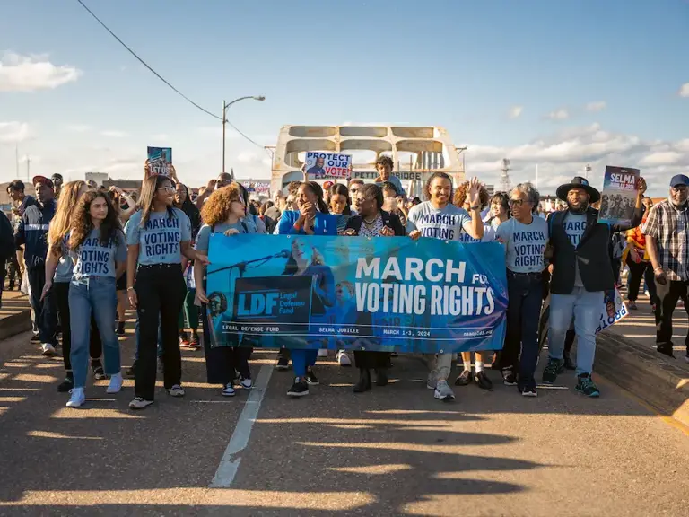 Color photo of LDF staff marching across the Edmund Pettus Bridge in Selma, Alabama, in March 2024 during the annual Selma Jubilee commemorating the anniversary of Bloody Sunday. Several people wear blue shirts and hold a banner reading, "March for Voting Rights."