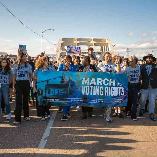 Color photo of LDF staff marching across the Edmund Pettus Bridge in Selma, Alabama, in March 2024 during the annual Selma Jubilee commemorating the anniversary of Bloody Sunday. Several people wear blue shirts and hold a banner reading, "March for Voting Rights."