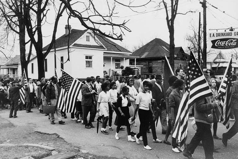 Black-and-white photo of activists, some carrying American flags, participating in the civil rights march from Selma to Montgomery, Alabama, in 1965.