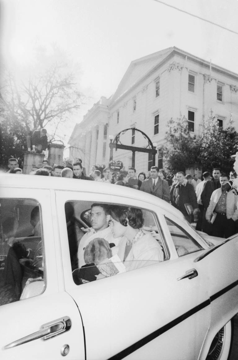 Black-and-white photo of Black students Charlayne Hunter and Hamilton Holmes leaving the campus of the University of Georgia in a car after they registered for classes on January 9, 1961, as a crowd of people look on.