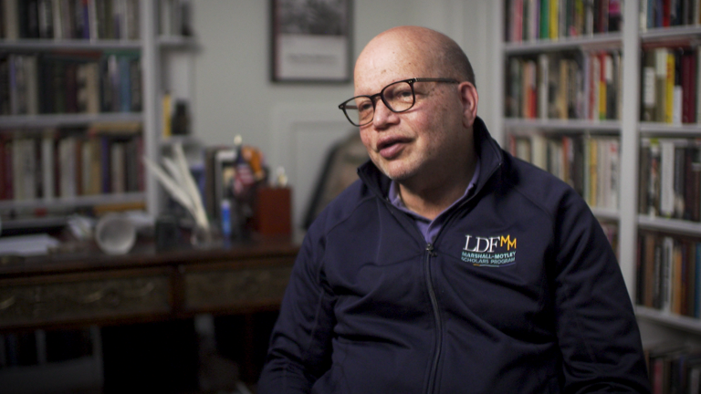 Color photo of Ted Shaw, wearing glasses and an LDF sweatshirt, sitting in front of bookshelves during his oral history interview.