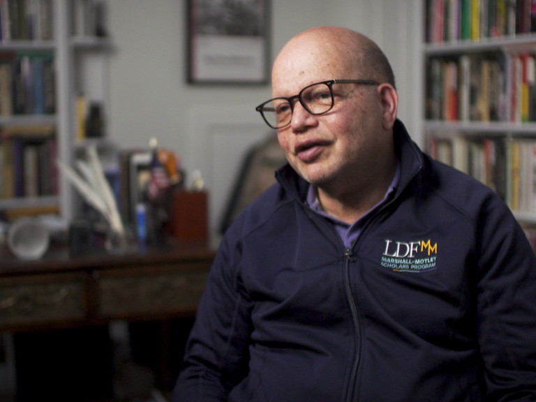 Color photo of Ted Shaw, wearing glasses and an LDF sweatshirt, sitting in front of bookshelves during his oral history interview.