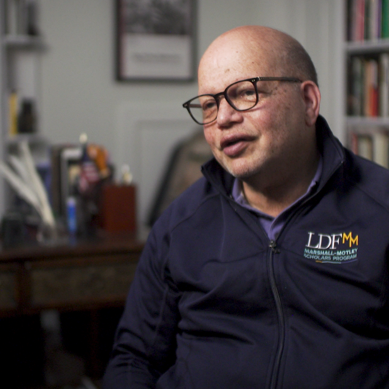 Color photo of Ted Shaw, wearing glasses and an LDF sweatshirt, sitting in front of bookshelves during his oral history interview.