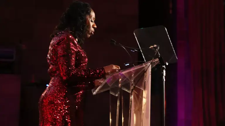 Color photo of Sherrilyn Ifill, wearing a shiny red dress, speaking at a podium indoors.