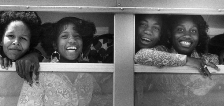 Black-and-white photo of four Black children smiling through a school bus window. In the early days of desegregation in Jefferson County, Kentucky, these students were bused between Jeffersontown and Central High School. 