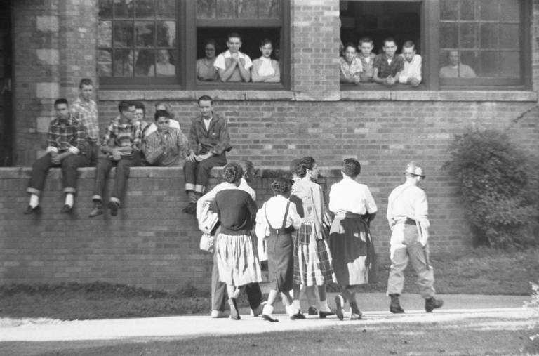 Black-and-white photo of white students sitting on a ledge and standing at school windows as they watch a group of Black students be escorted.