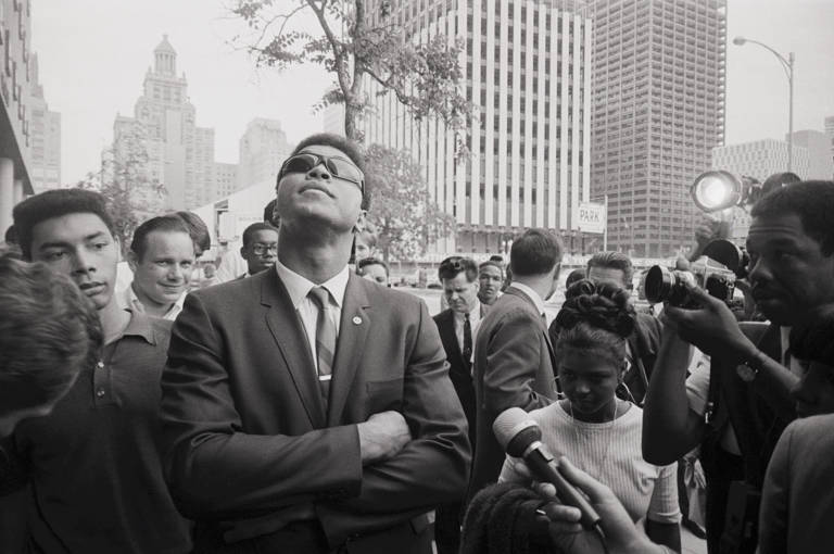 Black-and-white photo of boxing heavyweight champion Muhammad Ali looking skyward, surrounded by reporters, as he leaves federal court in Houston, Texas, after a federal judge tossed out his last legal effort to avoid being drafted into the Army on April 27, 1967.