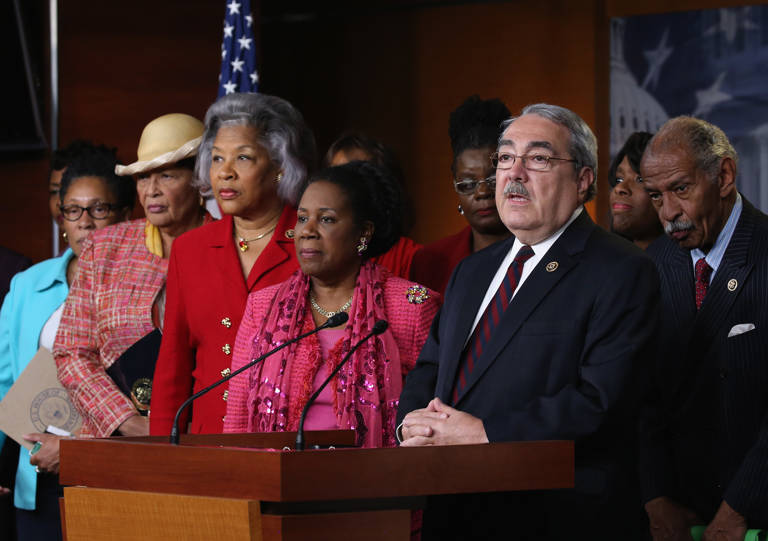 Color photo of U.S. Rep. G.K. Butterfield (right) wearing a suit and speaking behind a podium while flanked by members of the Congressional Black Caucus during a news conference on Capitol Hill on April 22, 2015.