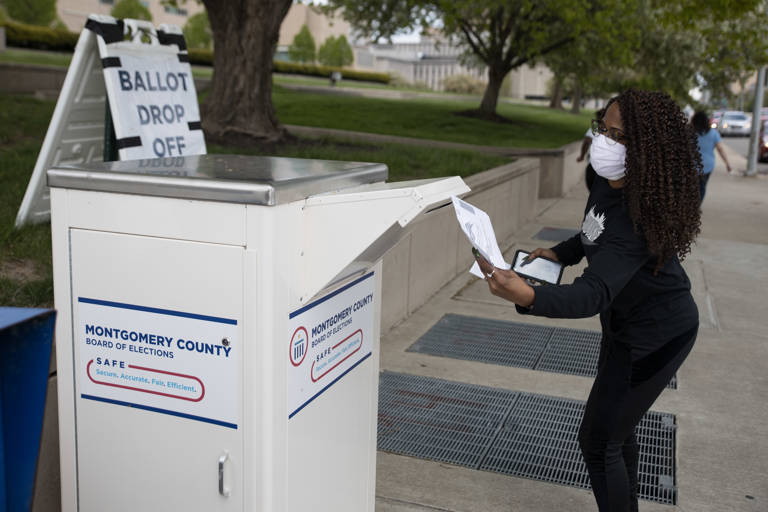 Color photo of a voter wearing a surgical mask dropping off a ballot in a large white container reading, "Montgomery County Board of Elections SAFE Secure, Accurate, Fair, and Efficient."