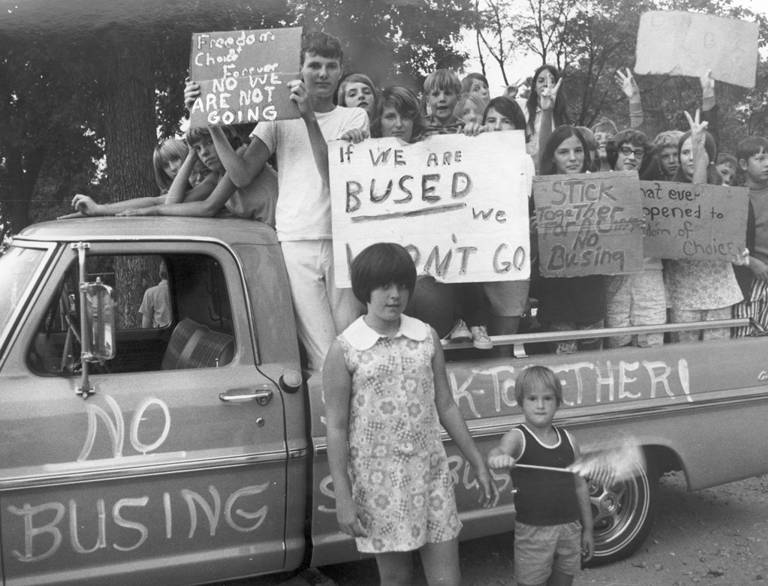 Black-and-white photo of a protest, with white children standing on a truck that has writing on the side stating, "no busing" and "stick together." The children are holding signs stating, "Freedom of choice forever, no we are not going," "If we are bused we won’t go," and "Stick together for a change, no busing."