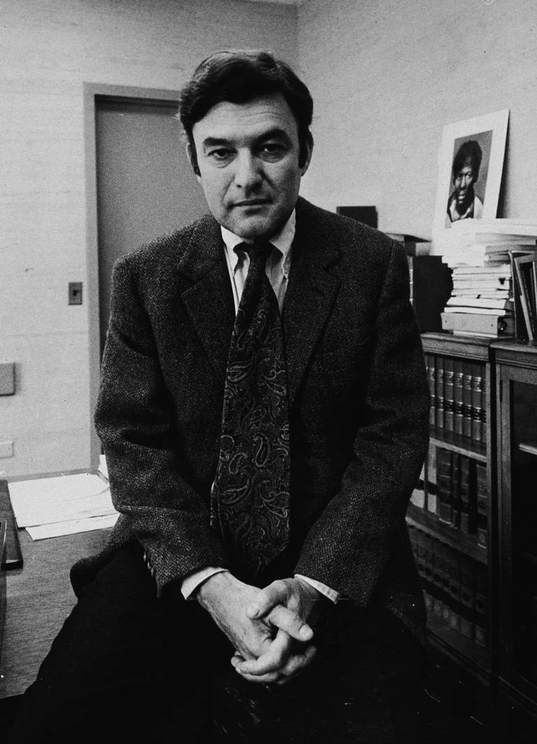 Black-and-white photo of Jack Greenberg sitting at the edge of his desk and wearing a suit. In the background are a bookcase and a photo.