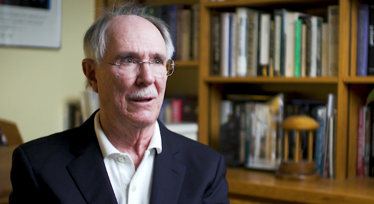 Color photo of Jack Boger, wearing a suit and sitting in front of a bookshelf during his oral history interview.