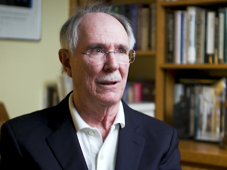 Color photo of Jack Boger, wearing a suit and sitting in front of a bookshelf during his oral history interview.