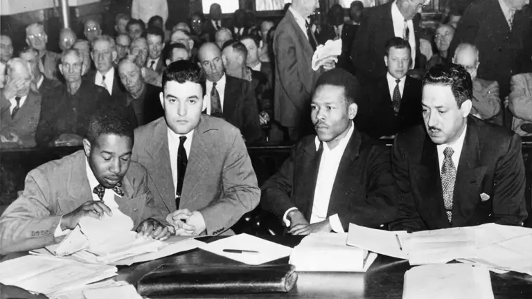 Black-and-white photo of four men wearing suits, including Jack Greenberg and Thurgood Marshall, seated behind a table covered in papers in a courtroom in front of a crowd of spectators.