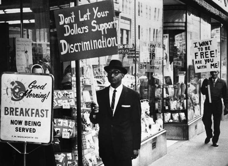 Black-and-white photo of picketers demanding an end to lunch counter segregation while marching outside a Woolworth store in Lynchburg, Virginia. The two picketers hold signs reading, "Don't Let Your Dollars Support Discrimination" and "I Want to Be Free! Are You With Me?"