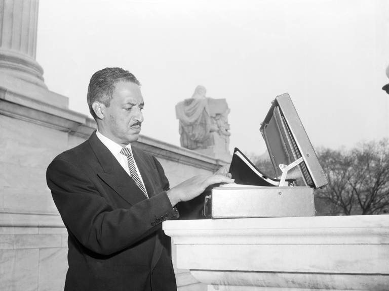 Black-and-white photo of Thurgood Marshall wearing a suit and tie as he pauses to organize his papers in an open briefcase outside the U.S. Supreme Court building. 