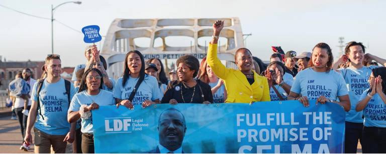 Color photo of LDF staff, including President and Director-Counsel Janai Nelson and Associate Director-Counsel Tona Boyd, marching across the Edmund Pettus Bridge while holding a banner that reads, "Fulfill the Promise of Selma."