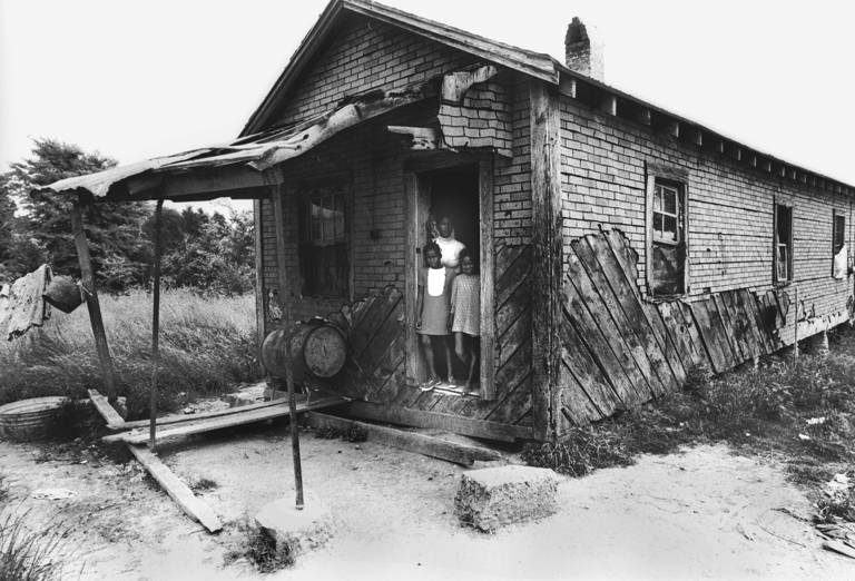 Black-and-white photo of a mother standing in the doorway of a dilapidated plantation farmhouse with her two daughters, somewhere in the Mississippi Delta territories in 1971.