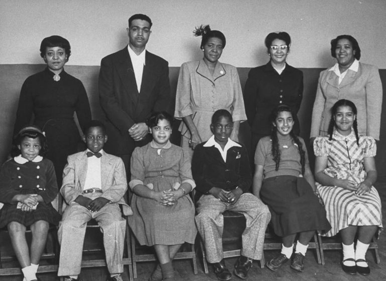 Black-and-white photo of six Black children sitting in a row and five Black adults standing behind them, all wearing suits and dresses.