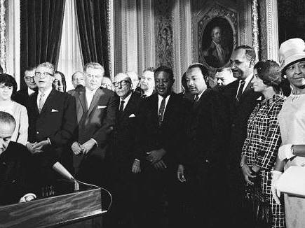 Black-and-white photo of President Lyndon B. Johnson sitting behind a podium as a large group of men and women stand and watch him.