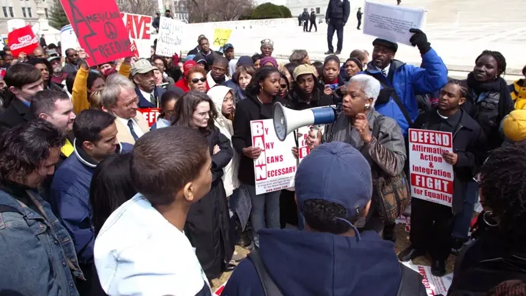 Color photo of Elaine Jones holding a bullhorn and speaking to a large crowd of demonstrators, who hold signs reading, "American Freedom," "By Any Means Necessary," and "Defend Affirmative Action and Integration, Fight for Equality."