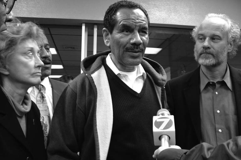 Black-and-white photo of Wilbert Rideau standing between LDF counsel as he speaks to the press through a microphone.