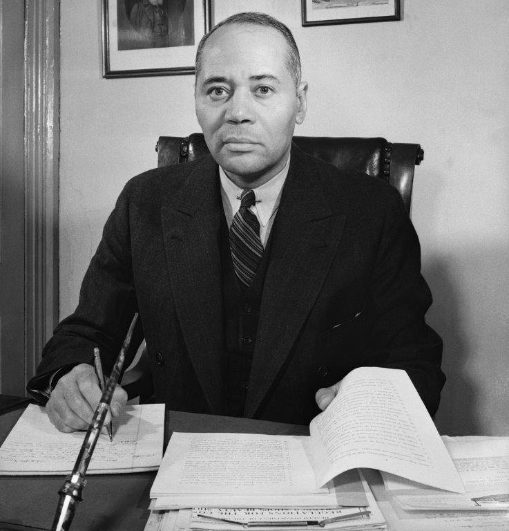 Black-and-white photo of NAACP General Counsel Charles Hamilton Houston sitting at his desk and writing in a notebook.