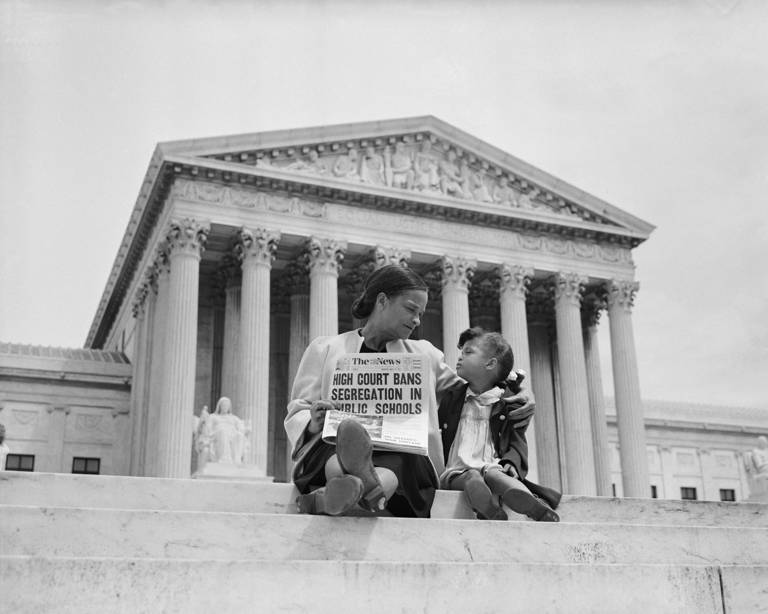 Black-and-white photo of Nettie Hunt and her daughter Nickie sitting on the steps of the U.S. Supreme Court building, as Nettie holds a newspaper with a headline reading, "High Court Bans Segregation in Public Schools."