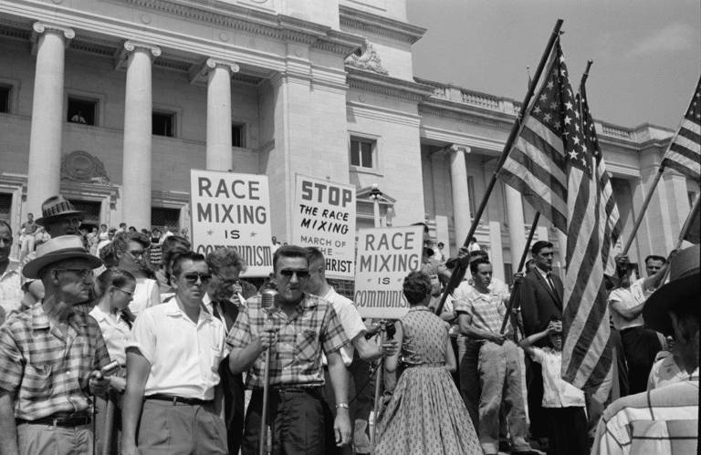 Photo of people gathered at the Arkansas state capitol building, several holding signs and American flags, to protest the admission of the "Little Rock Nine" to Central High School. 