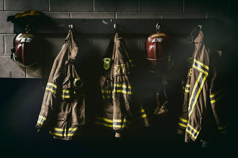 Color photo of firefighter suits and helmets hanging in a dark room.