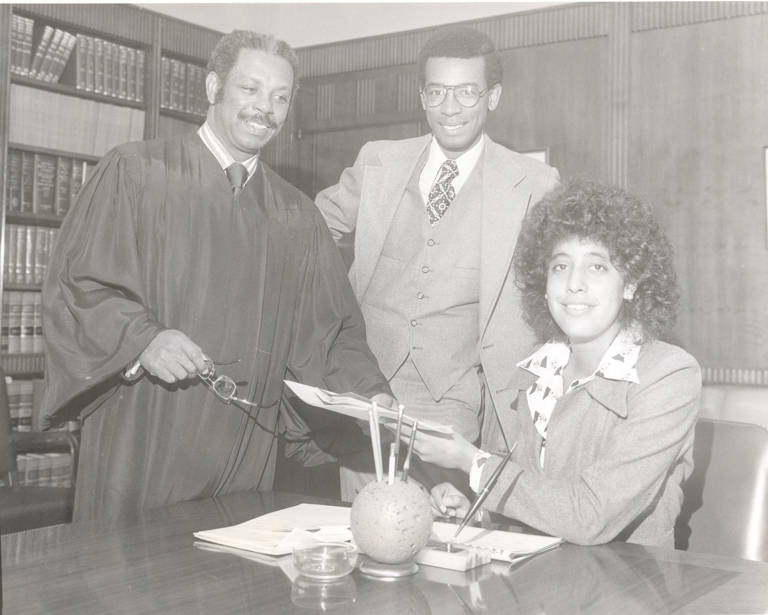 Black-and-white photo of Lani Guinier (sitting, wearing a suit, and holding papers, right) with the Honorable Damon J. Keith (standing, in a black robe, far left) and James Coleman (standing, in a suit, middle). 