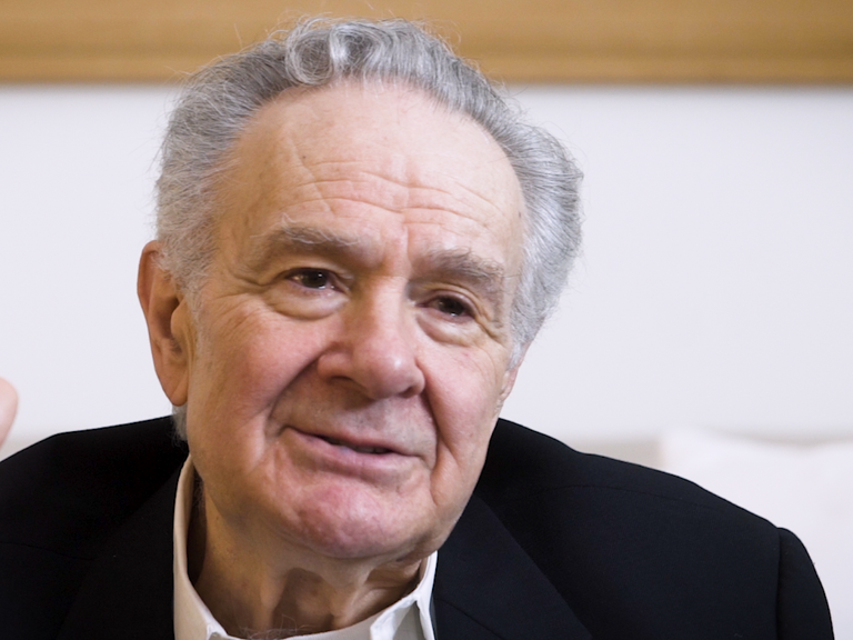 Color photo of Melvyn Leventhal, seated indoors with his hand raised, during his oral history interview.