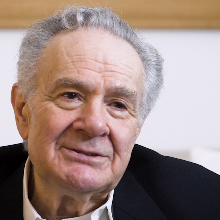 Color photo of Melvyn Leventhal, seated indoors with his hand raised, during his oral history interview.