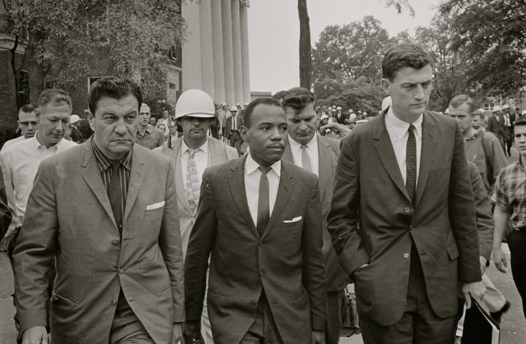 Black-and-white photo of James Meredith walking to class, wearing a suit and tie, accompanied by U.S. marshals and lawyer John Doar (right) in 1962.