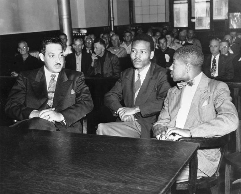 Black-and-white photo of three men wearing suits (from left to right, Thurgood Marshall, Walter Lee Irvin, and Paul C. Perkins Jr.) sitting behind a table in a courtroom, with a crowd of seated spectators behind them.