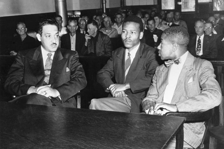 Black-and-white photo of three men wearing suits (from left to right, Thurgood Marshall, Walter Lee Irvin, and Paul C. Perkins Jr.) sitting behind a table in a courtroom, with a crowd of seated spectators behind them.