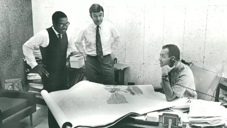 Black-and-white photo of Julius Levonne Chambers and Jack Greenberg, standing behind a desk with several papers and maps, while a third man is seated behind the desk talking on the phone.