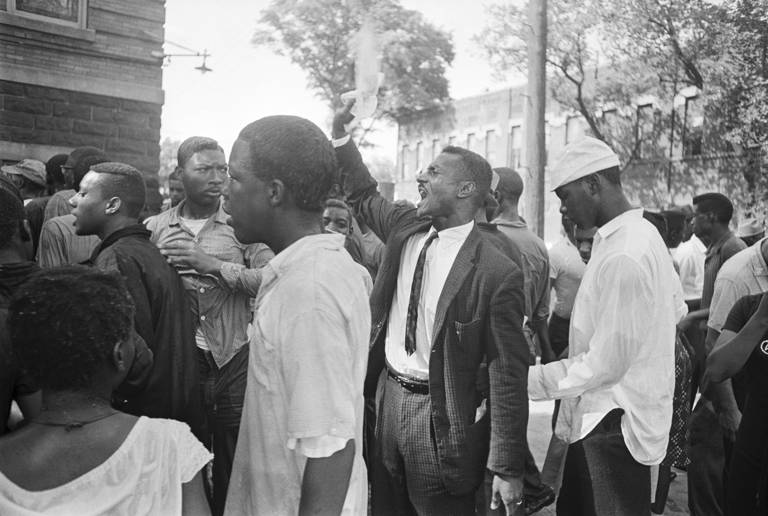 Black-and-white photo of the Rev. Fred Shuttlesworth, wearing a suit and tie and with his hand raised, speaking to a group of Black protesters after a segregation protest was disrupted by people throwing rocks and bottles in Birmingham, Alabama, on May 8, 1963.